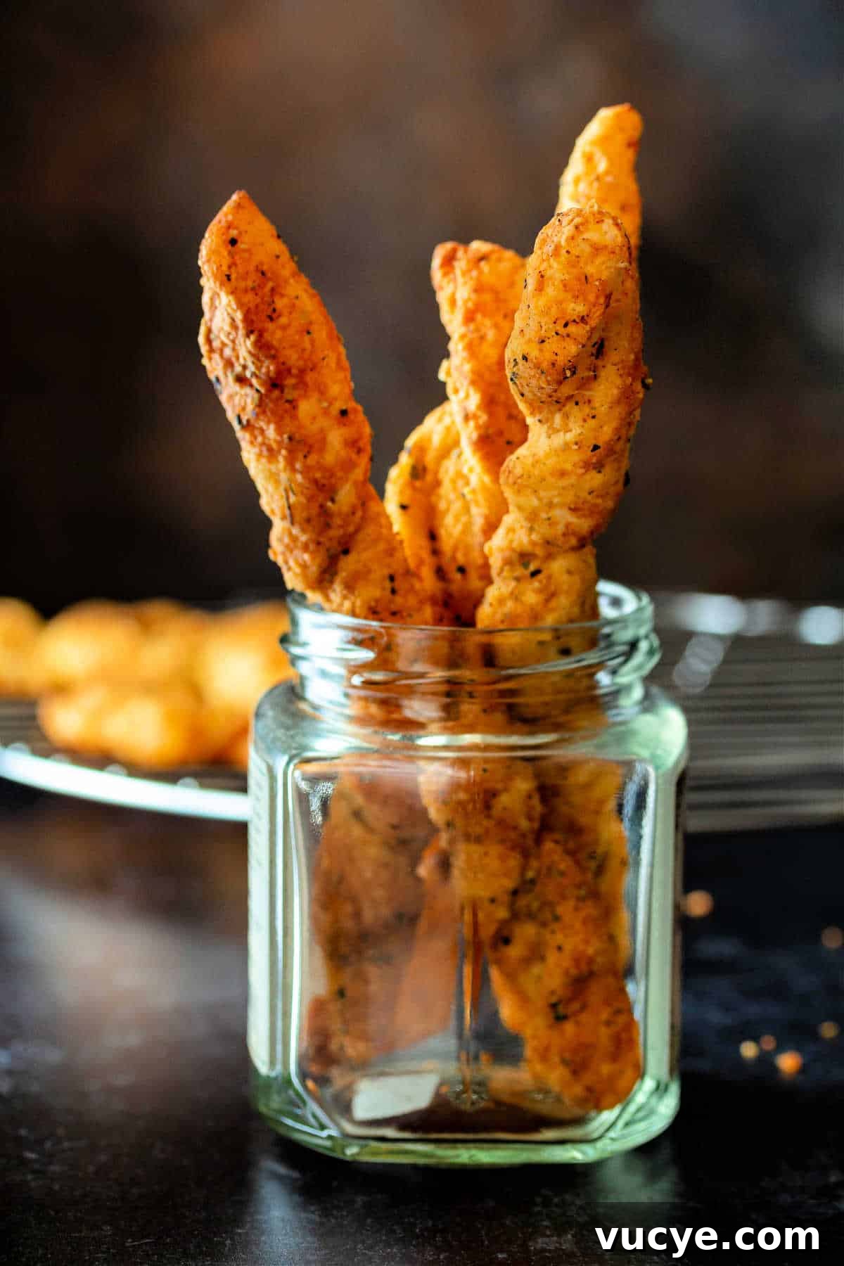 Crispy Puff Pastry Cheese Straws Recipe 2 A glass jar with cheese straws in it with more cooling in the background on a metal cooling rack.