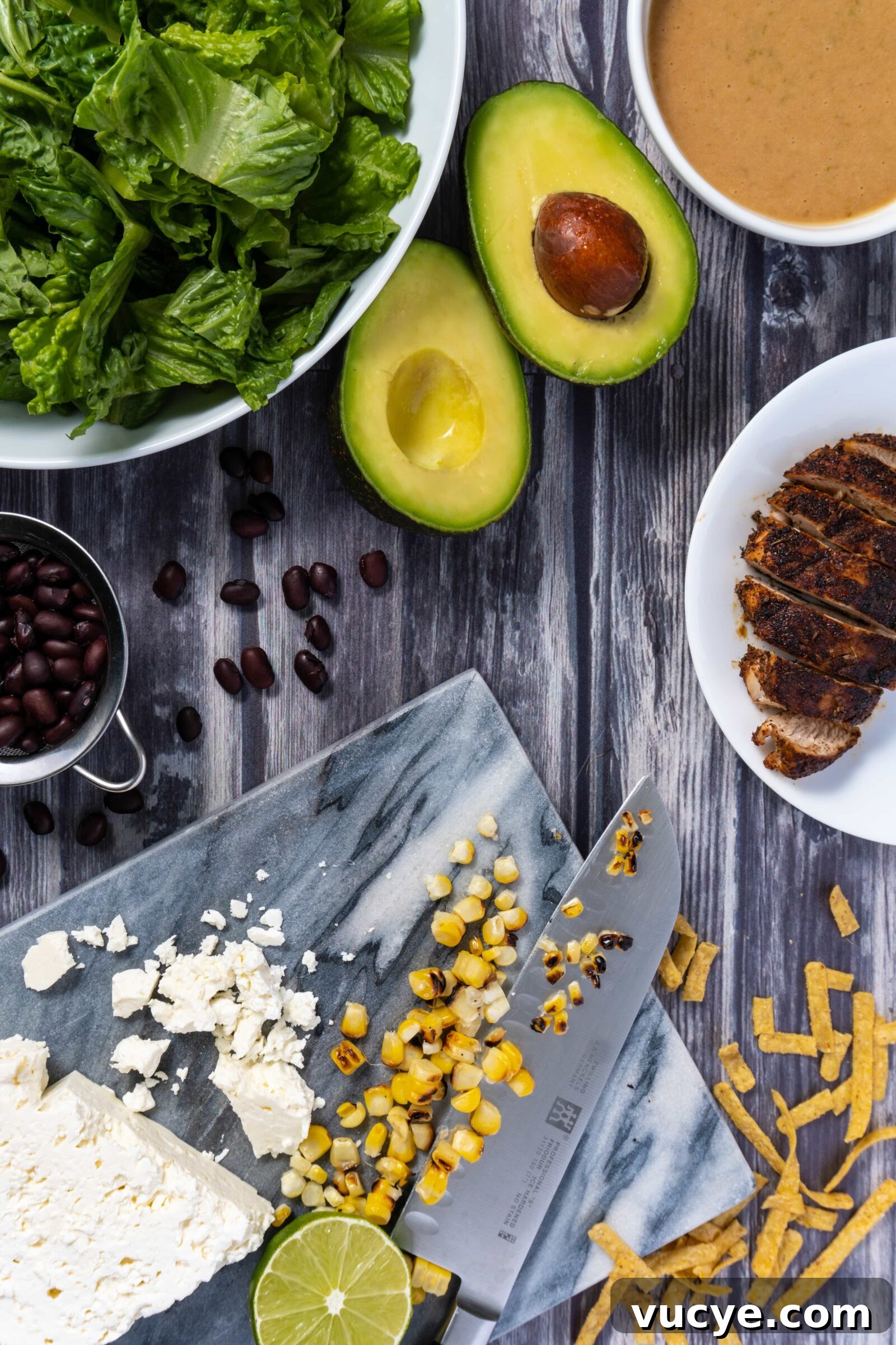 Cajun chicken, romaine lettuce, corn. feta, black beans, dates, tortilla strips, avocado, and limes are ready to be added into a bowl and tossed with peanut lime vinaigrette