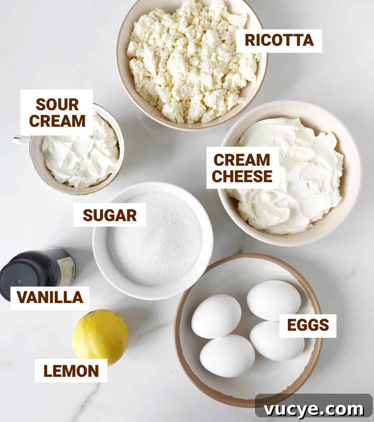 Silky Ricotta Cheesecake Recipe for a Light, Creamy Dessert 4 Ingredients for ricotta cheesecake in bowls on white marble, including lemon, vanilla, sugar, eggs, sour cream, cream cheese.