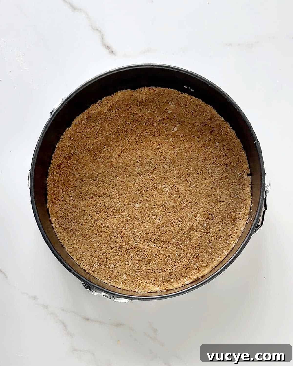 Silky Ricotta Cheesecake Recipe for a Light, Creamy Dessert 5 Graham cracker crust for cheesecake in a round dark metal cake pan on white marble.