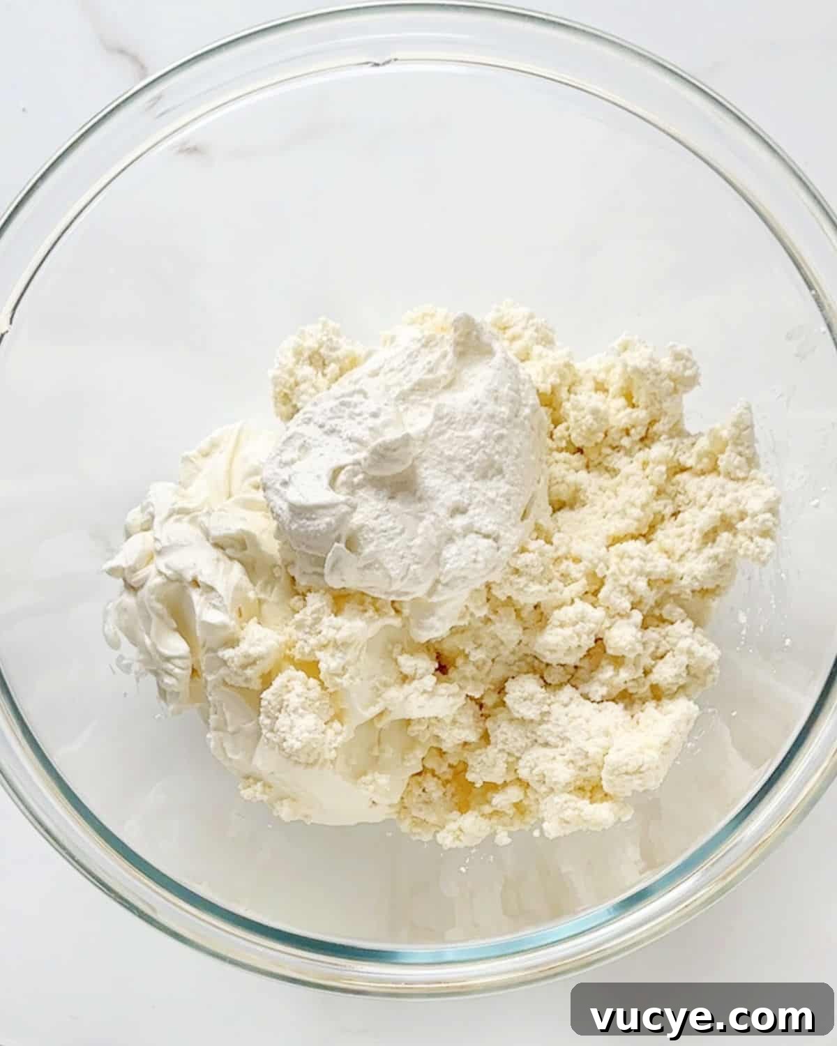 Silky Ricotta Cheesecake Recipe for a Light, Creamy Dessert 6 Glass bowl with ricotta, cream cheese, and sour cream. White marble surface.
