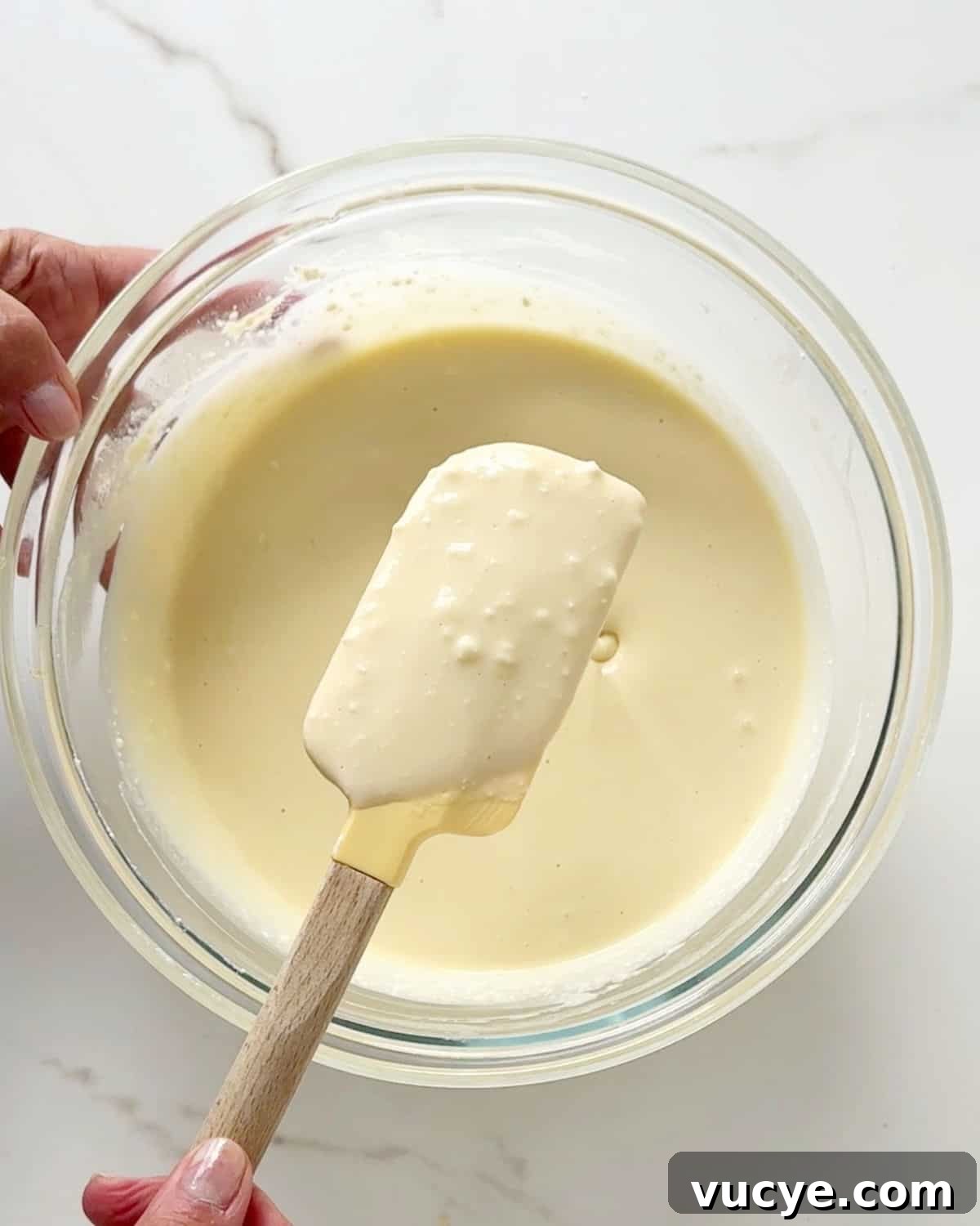 Silky Ricotta Cheesecake Recipe for a Light, Creamy Dessert 7 Ricotta cheesecake batter texture being shown with a spatula from a glass bowl. White marble surface.