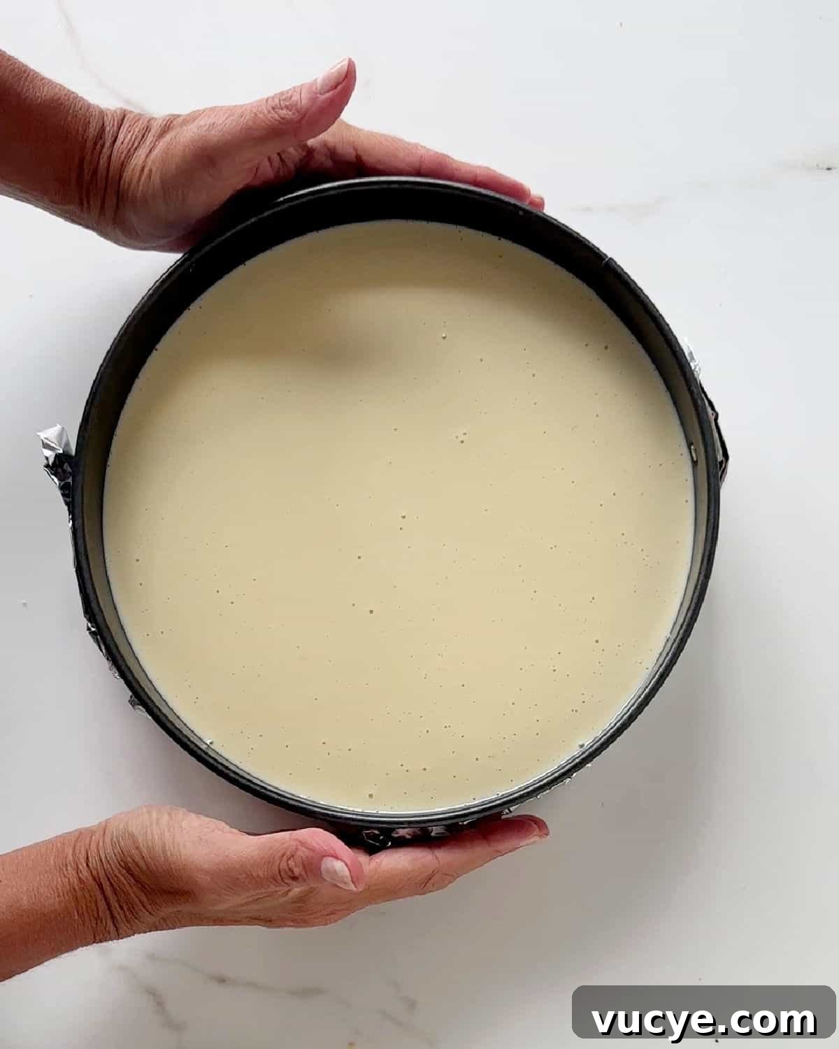 Silky Ricotta Cheesecake Recipe for a Light, Creamy Dessert 8 Hands holding a round cake pan with ricotta cheesecake filling before baking. White marble surface.
