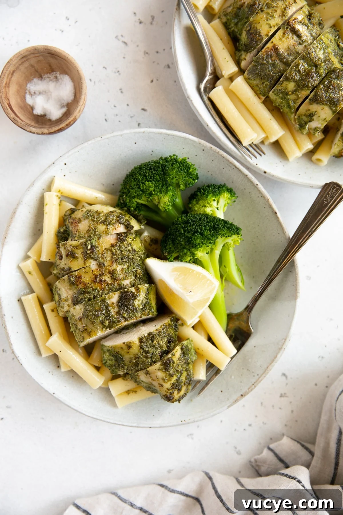 One-Pan Pesto Chicken with Roasted Vegetables 8 Baked pesto chicken recipe on a white serving plate with cooked pasta noodles, fresh lemon wedges, and steamed broccoli.