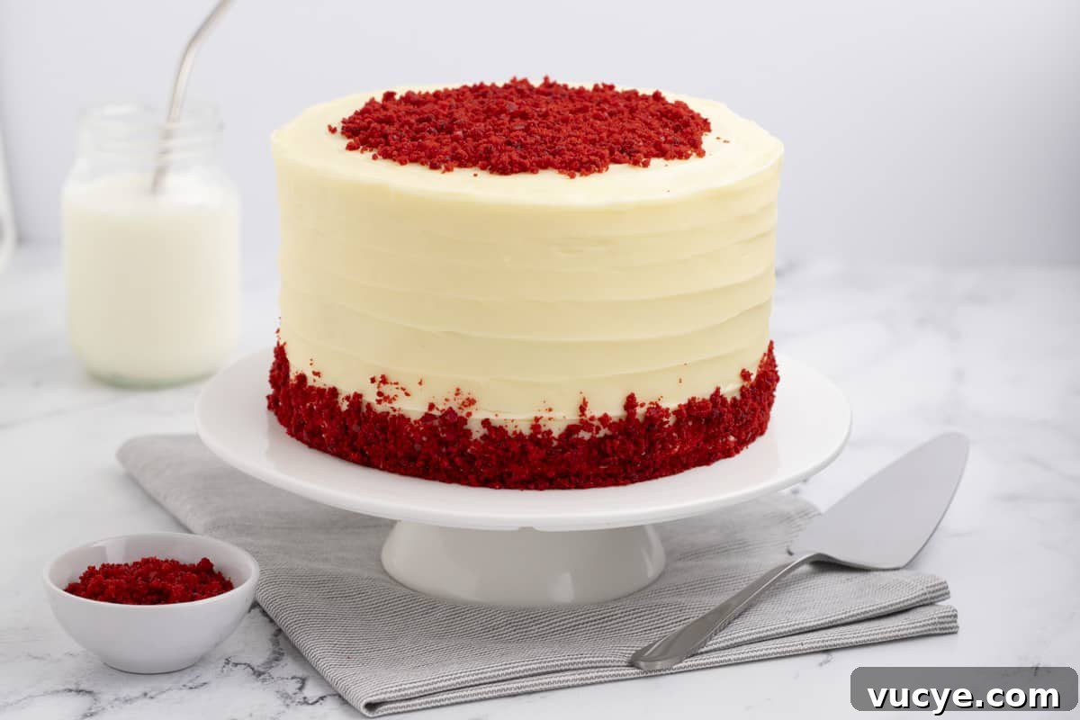 Homemade Red Velvet Cake Recipe From Scratch 2 red velvet cake with cream cheese frosting on a white cake stand with a bowl of cake crumbs, a serving knife and glass of milk beside it