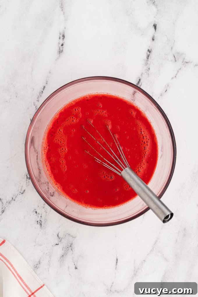 Homemade Red Velvet Cake Recipe From Scratch 11 wet ingredients with bright red food coloring in it in a clear glass bowl with a wire whisk
