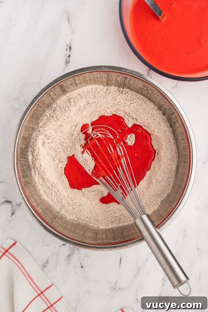Homemade Red Velvet Cake Recipe From Scratch 12 dry ingredients for red velvet cake with red wet ingredients being poured in and mixed with a wire whisk in a silver bowl