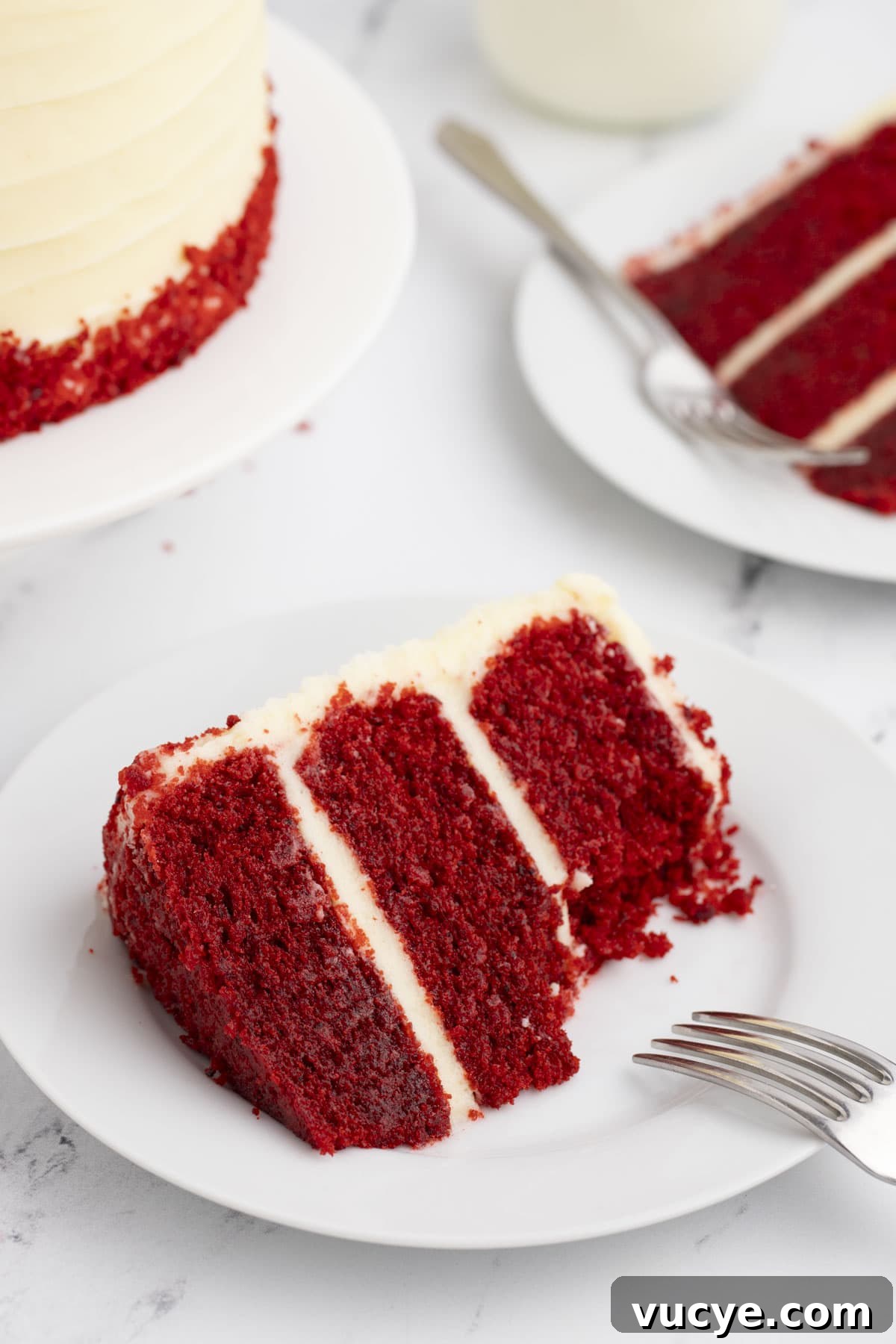 Homemade Red Velvet Cake Recipe From Scratch 3 a slice of layered red velvet cake on a white plate with a fork leaning on it and another plate with a slice of cake and the edge of the whole cake in the background