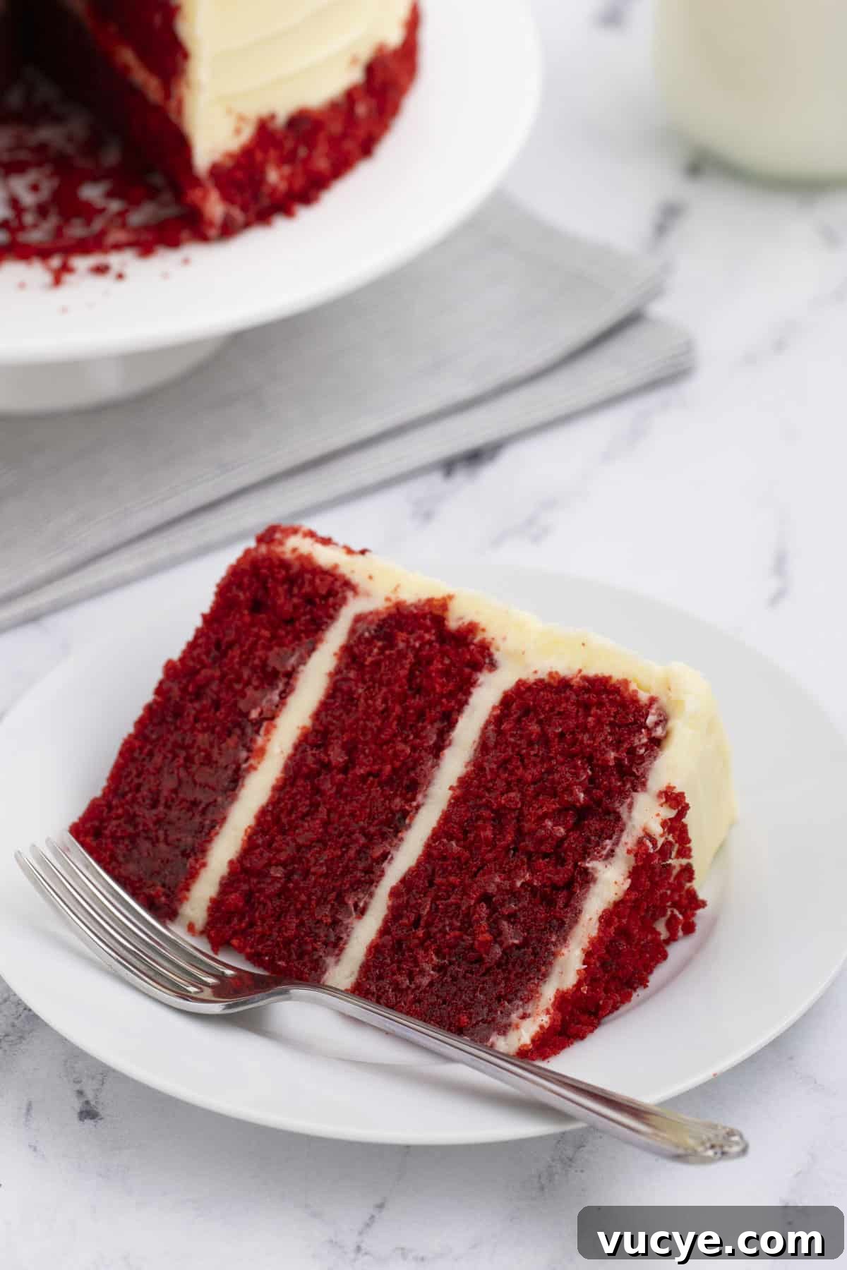 Homemade Red Velvet Cake Recipe From Scratch 15 a slice of red velvet cake on a small white plate with a fork with the rest of the cake behind it on a white plate on top of a grey towel