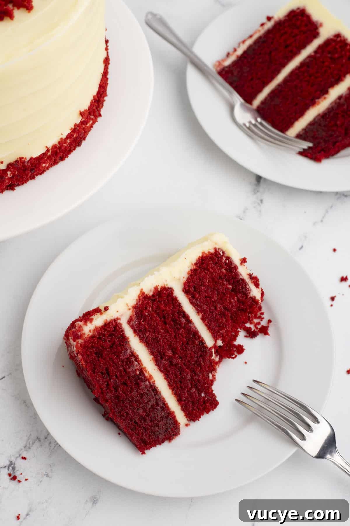 Homemade Red Velvet Cake Recipe From Scratch 6 a slice of layered red velvet cake on a white plate with a fork leaning on it and another plate with a slice of cake and the edge of the whole cake in the background