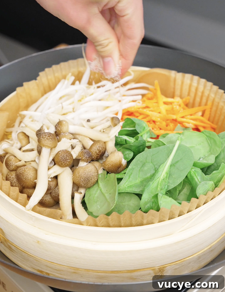 Authentic Beef Bibimbap Recipe with Crispy Veggies and Gochujang 5 Hand sprinkling salt over mushrooms bean sprouts spinach and carrots in a steamer for beef bibimbap recipe.