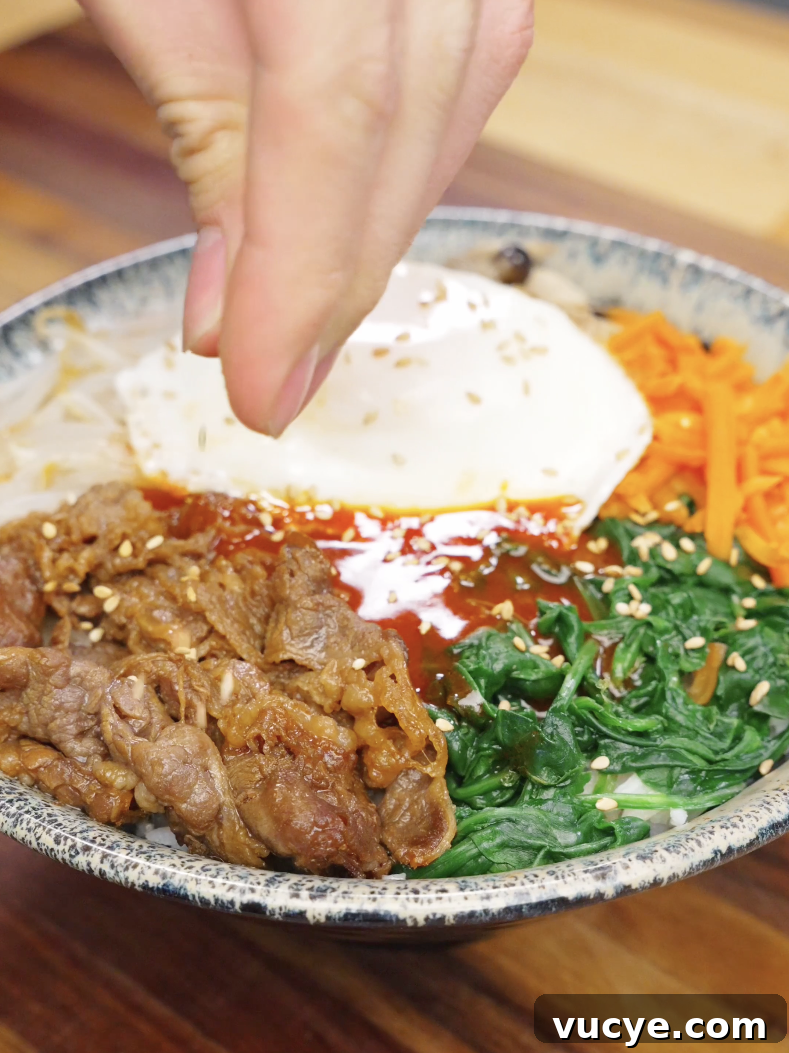 Authentic Beef Bibimbap Recipe with Crispy Veggies and Gochujang 8 Hand sprinkling sesame seeds over beef bibimbap recipe with rice vegetables and fried egg.
