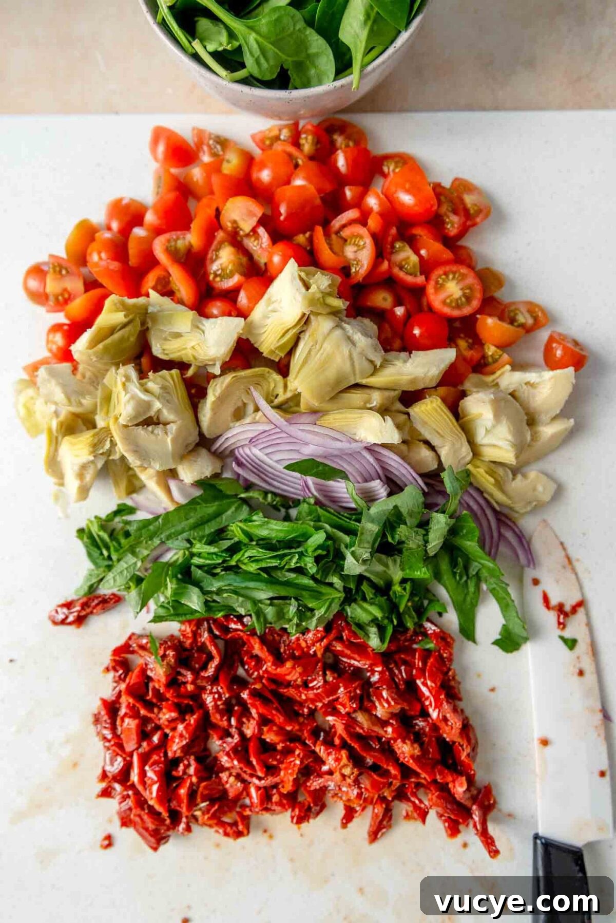 Chopped grape tomatoes, artichoke hearts, basil, red onion and sun dried tomatoes on a cutting board.