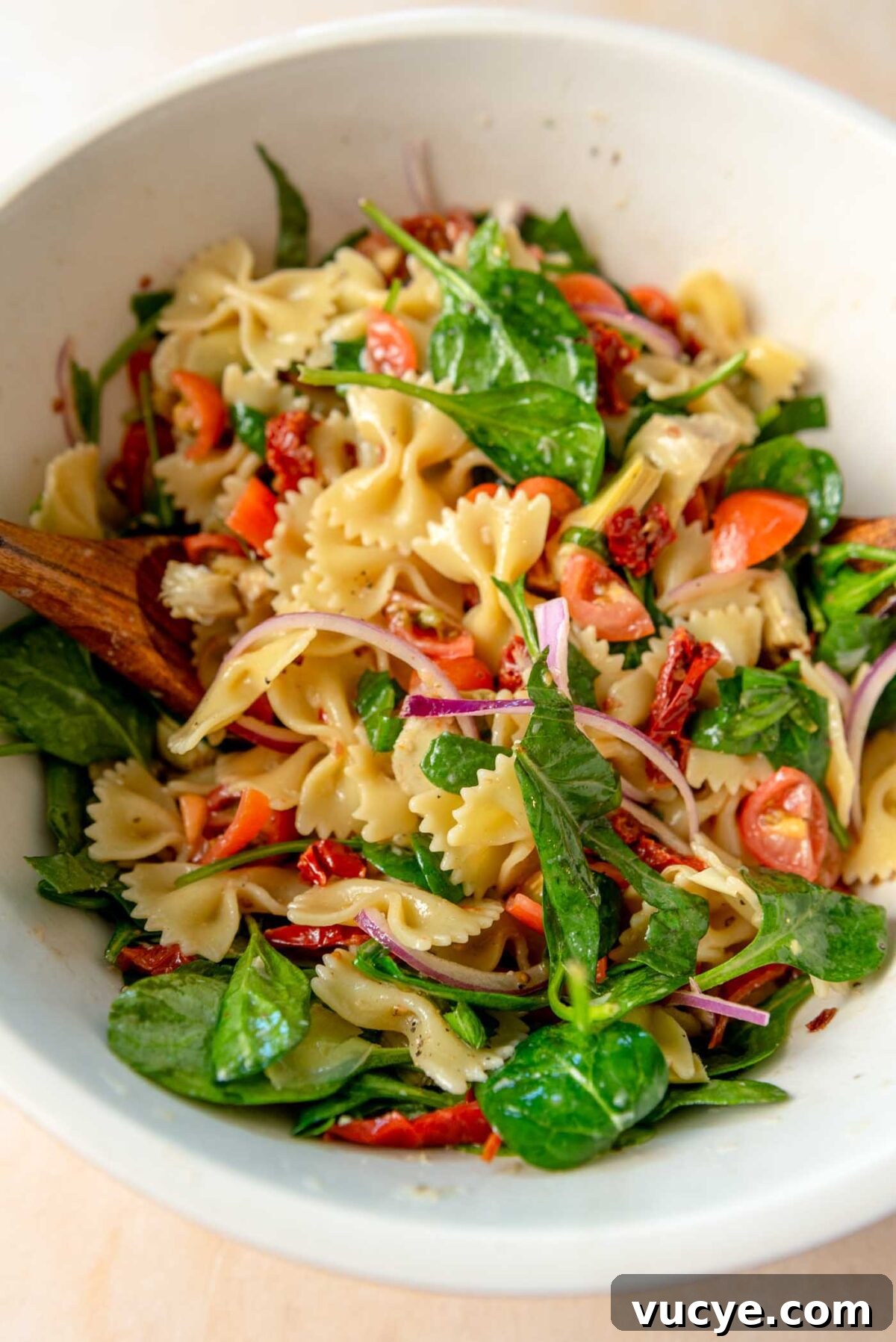 Sun dried tomato pasta salad with fresh spinach, red onion, cherry tomatoes and artichokes in a mixing bowl.
