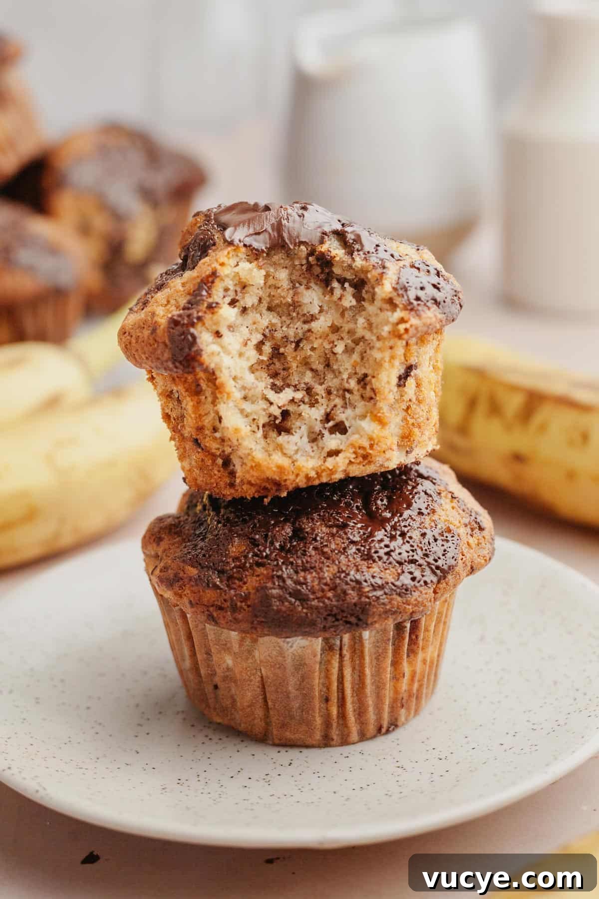 Bakery-Style Chocolate Chunk Banana Muffins Recipe 2 Two muffins stacked on top of each other, the muffin on top has a bite taken out of it.