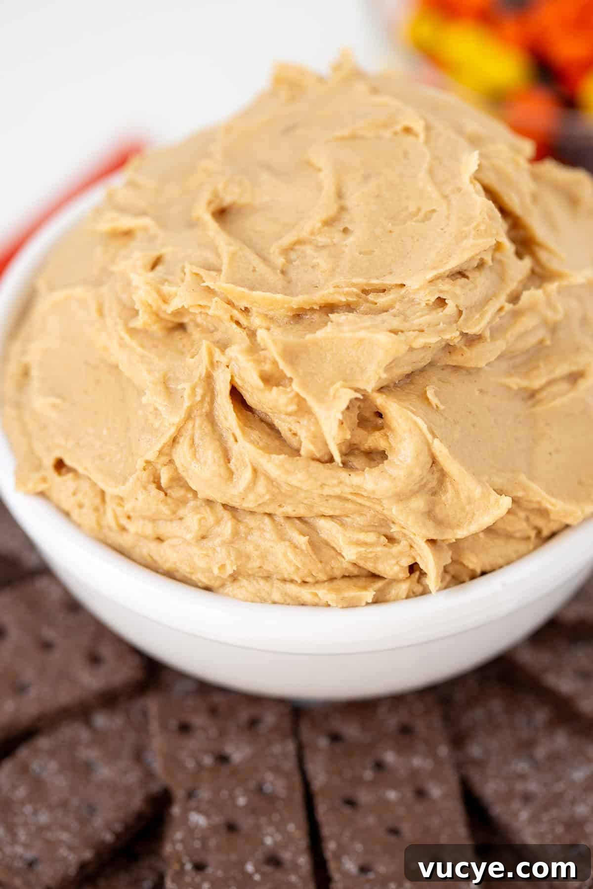 Peanut Butter Cookie Dough Dip Recipe — No-Bake Snack Dip 5 A white bowl filled with creamy Peanut Butter Dip sits next to chocolate cookies, with a blurred bowl of colorful candy in the background.