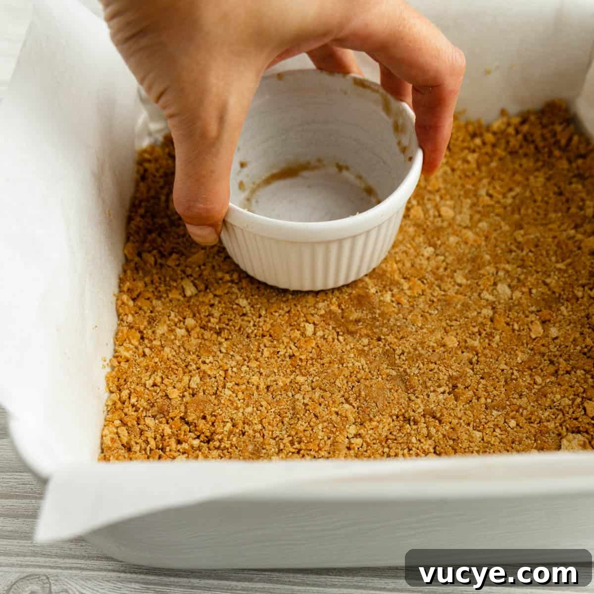 Creamy No-Bake Cheesecake Bars You Can Make Tonight 6 A hand holding a cup, pressing graham cracker crumbs into a pan.