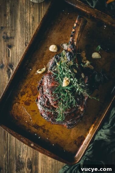 A cooked tomahawk steak garnished with herbs and garlic cloves rests on a baking sheet over a wooden table.