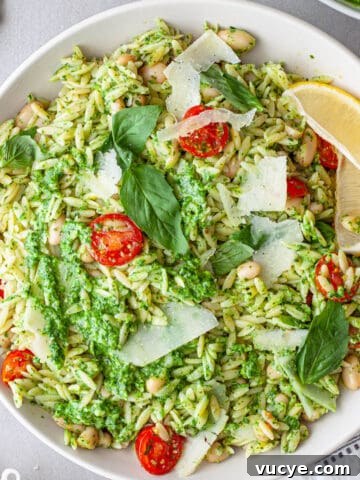 A white serving bowl of pesto pasta with chopped tomatoes, basil leaves and shaved parmesan on top.