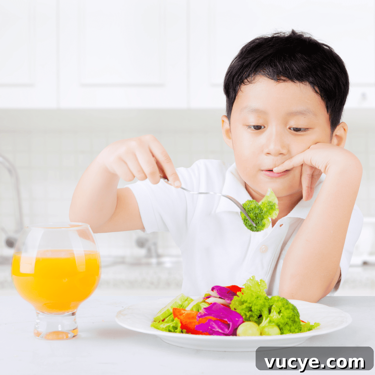 A boy in a white kitchen eating a colorful plate of vegetables with a clear glass of orange juice.
