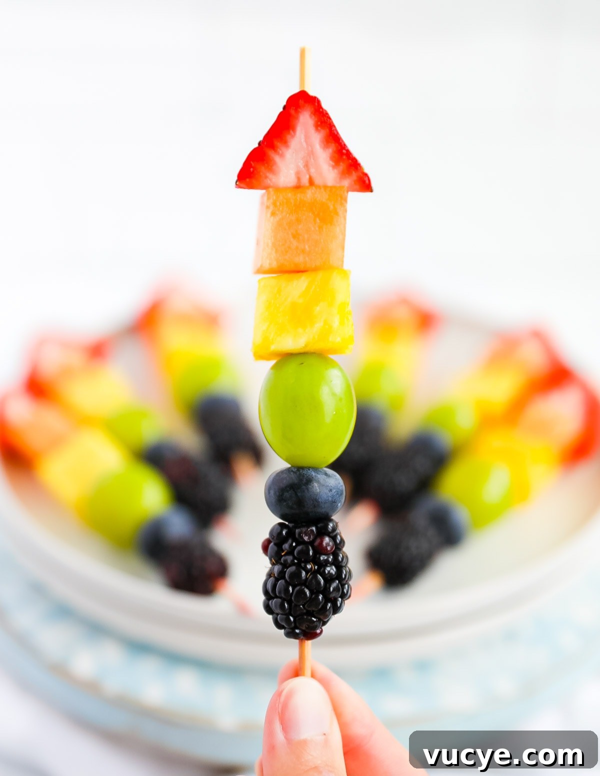 Colorful Rainbow Fruit Skewers Recipe for Parties and Kids 2 A hand holding up a wooden stick with strawberry, melon, pineapple, grape, blueberry, and blackberry on it