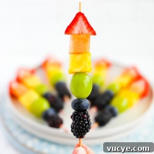 Colorful Rainbow Fruit Skewers Recipe for Parties and Kids 11 A hand holding up a wooden stick with one piece of strawberry, melon, pineapple, grape, blueberry, and blackberry