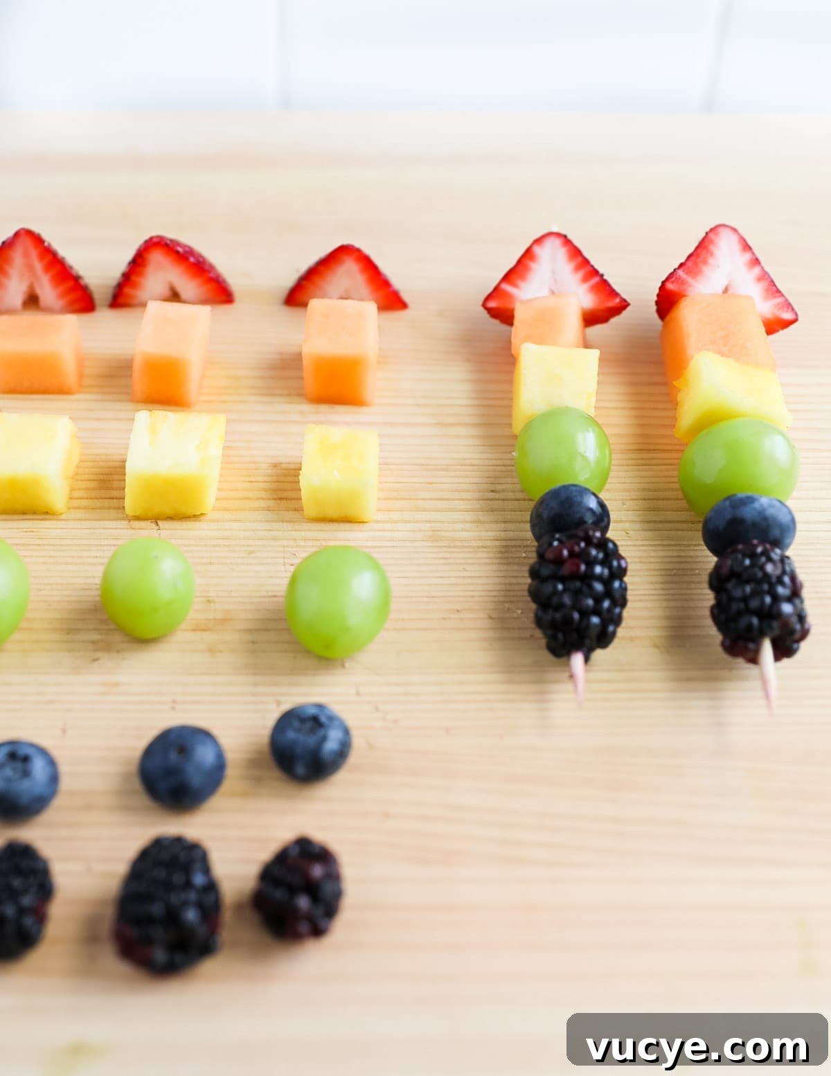 Colorful Rainbow Fruit Skewers Recipe for Parties and Kids 4 How to make rainbow fruit skewers: Strawberry, melon, pineapple, green grapes, blueberries, and blackberries on a cutting board waiting to be assembled on wooden sticks