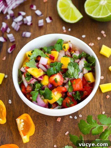 A white bowl sitting on a dark wooden cutting board, the bowl is filled with bright and colorful homemade mango salsa.