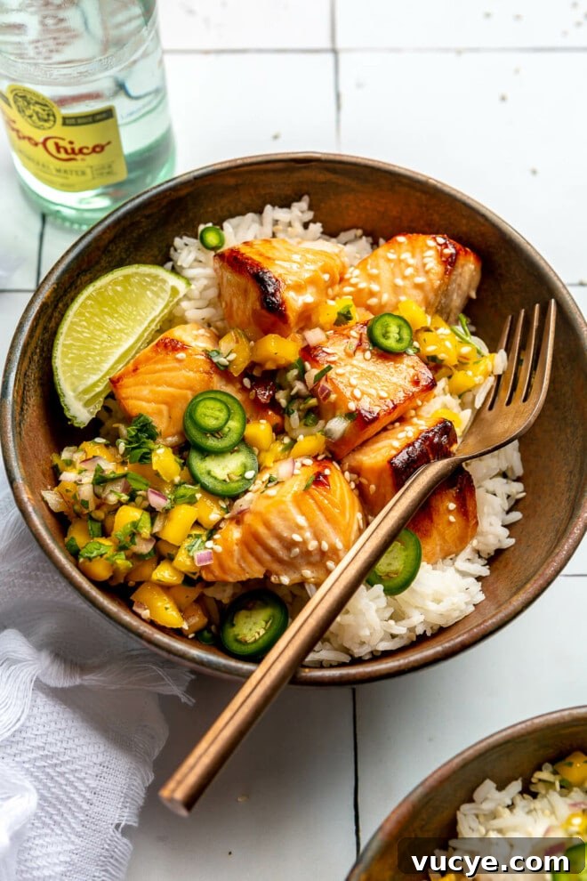 Maple Teriyaki Salmon Rice Bowls with Sesame & Scallions 4 White rice, teriyaki salmon, and mango salsa in a brown bowl sitting on a white tiled table next to a bottle of sparkling water and a white dish towel.