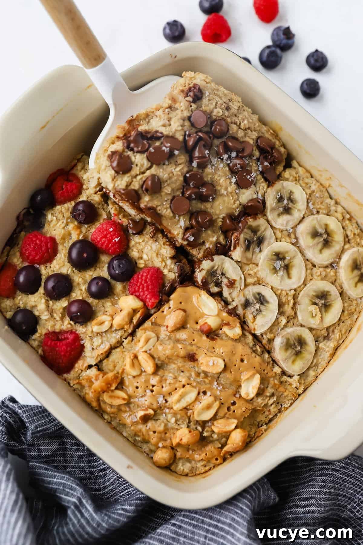 Chewy Baked Oatmeal Casserole with Berries and Cinnamon 2 A baking dish with four sections of baked oatmeal topped with chocolate chips, banana slices, mixed berries, and peanut butter with peanuts. A spatula rests on one section.