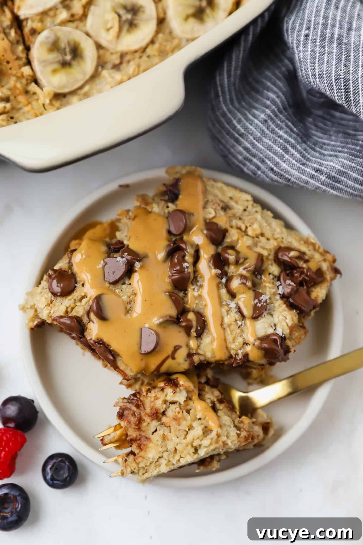 Chewy Baked Oatmeal Casserole with Berries and Cinnamon 6 A plate with a slice of banana baked oatmeal, freshly baked, topped with chocolate chips and a peanut butter drizzle. A fork rests on the plate, accompanied by vibrant blueberries and a raspberry on the side.