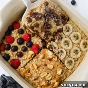 A dish of baked oatmeal is adorned with four tempting toppings: chocolate chips, banana slices, berries, and peanuts drizzled with peanut butter. A serving is being carefully lifted with a spatula, showcasing its delicious layers.