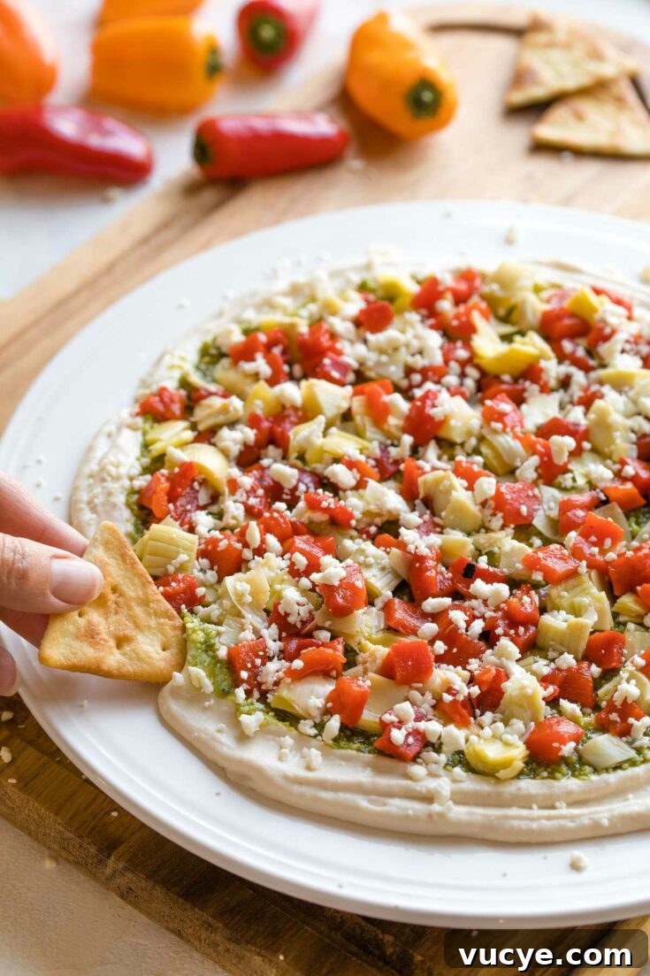 Hand dipping pita chip into layered pesto appetizer dip on white plate.