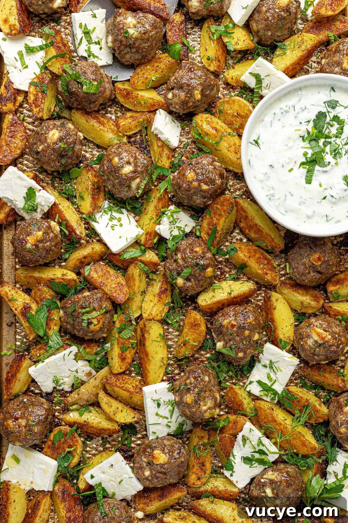 Greek-Style Sheet Pan Meatballs with Roasted Potatoes 10 Close up of a sheet pan filled with roasted potatoe, Greek meatballs, sliced feta, and yogurt sauce.
