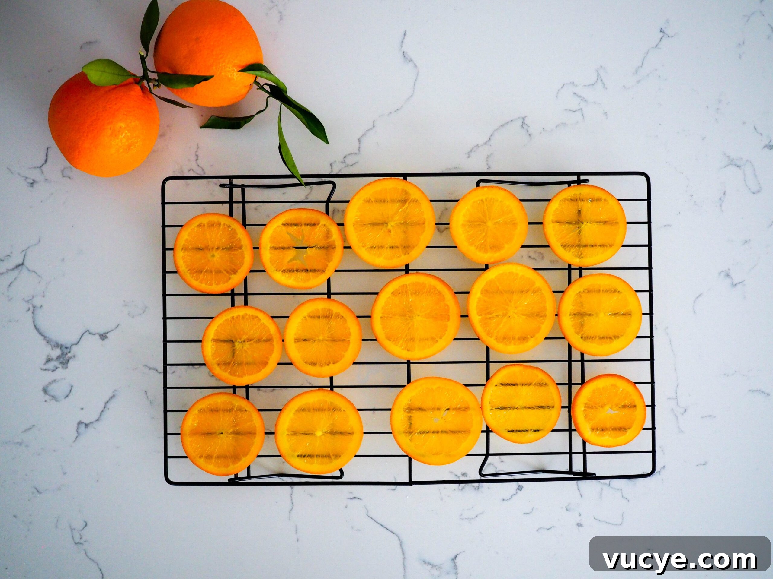 Candied Orange Slices Recipe: How to Make Sweet Orange Candy 5 Fifteen orange slices dry on a wire rack with two leafy oranges beside them.
