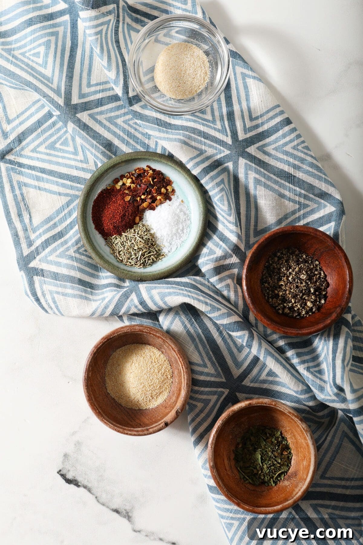 Ingredients to make a steak seasoning blend in bowls on a marble countertop with a blue patterned towel
