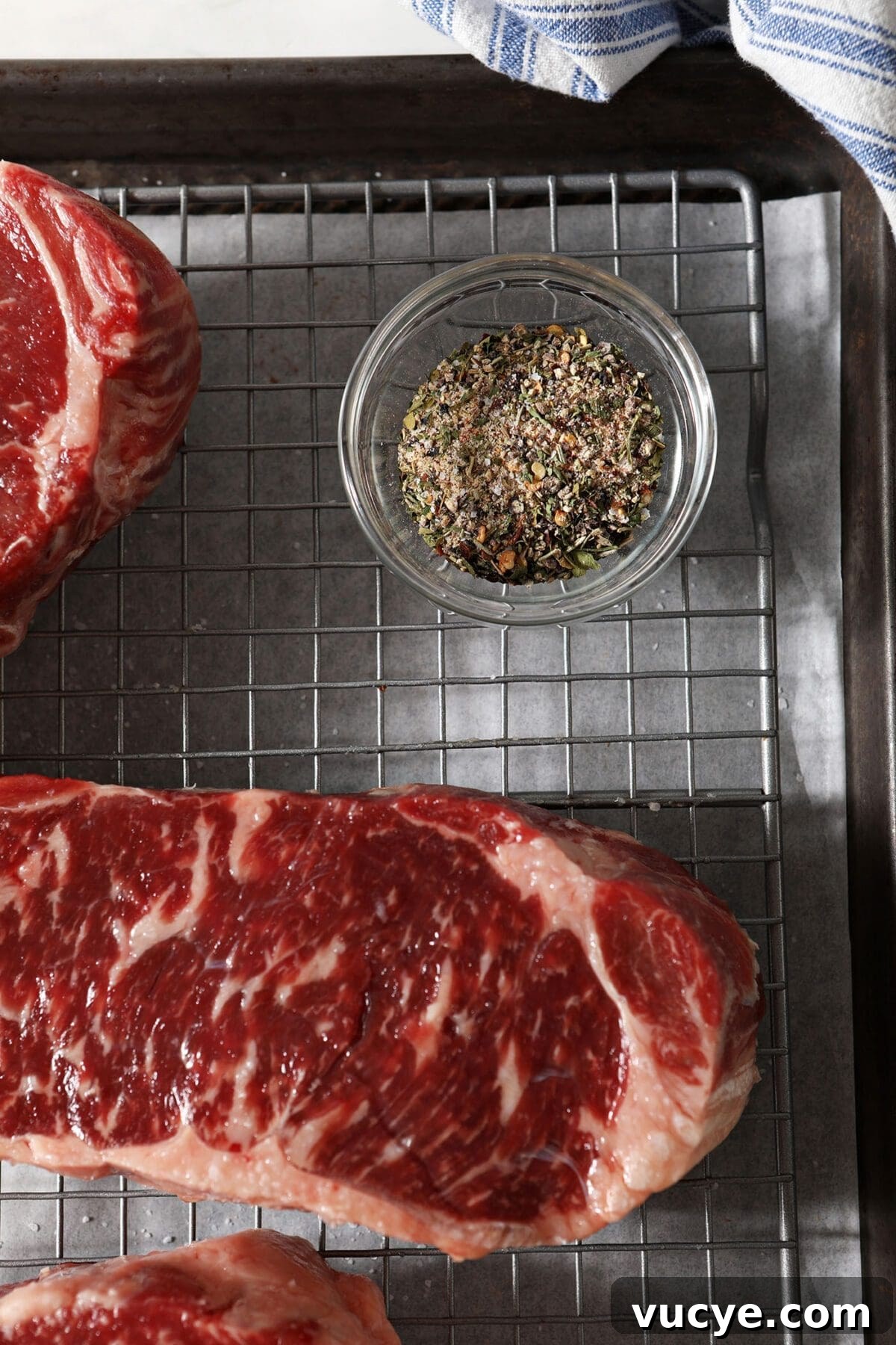 Steak seasoning blend in a small glass bowl next to steaks on a wire rack