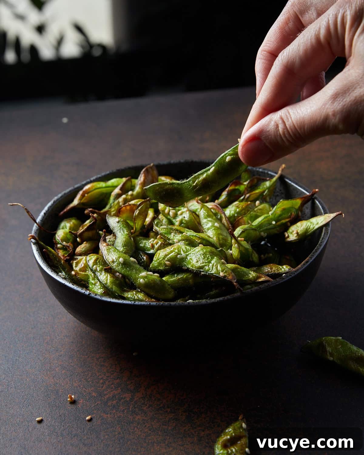 Crispy Air Fryer Frozen Edamame in 10 Minutes 5 Photo of a black bowl filled with air fryer frozen edamame beans set on a rusted black surface with a hand taking out one of the beans.