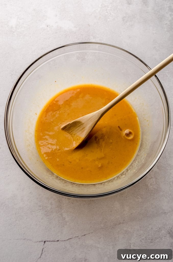 A mixture of pumpkin and other wet ingredients to make pumpkin cinnamon rolls in a large bowl with a wooden spoon.