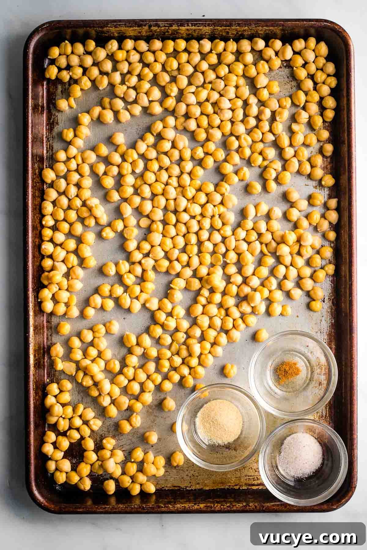 Chickpeas in a single layer on a baking sheet with three small dishes with spices.