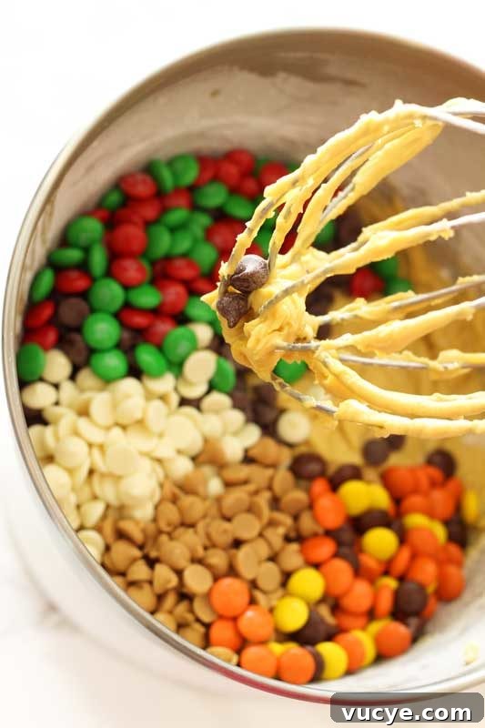 KitchenAid Standing Mixer containing a wire whisk with cookie dough; M&Ms, Reeses pieces and white and dark chocolate chips in a silver mixing bowl.