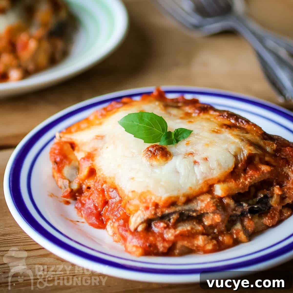 Side angled shot of eggplant polenta lasagna garnished with basil on a white and blue plate on a wooden table with silverware in the background.